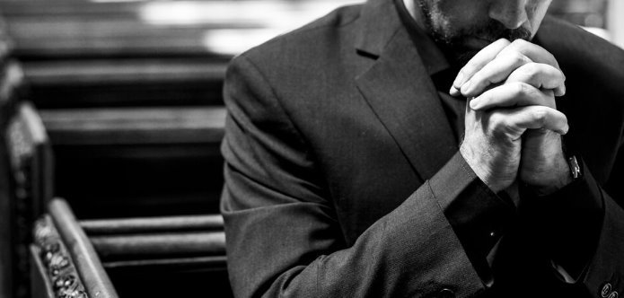 A man sitting in a church pew with his hands folded in prayer